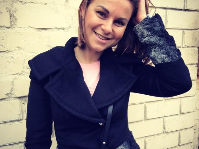 BelleLisaG with brown hair, smiling, wearing a black jacket with gray textured cuffs, pink shirt, hand in hair, standing against a white brick wall.