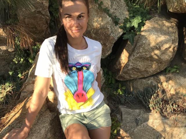 BelleLisaG com rabo de cavalo castanho, camiseta branca com rosto de macaco-azul e amarelo colorido, shorts verdes, sentada em pedras, sorrindo, com a luz do sol projetando sombras..