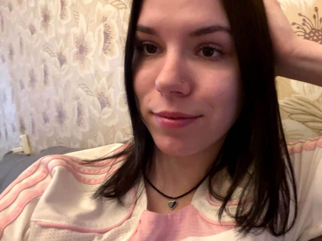 MagdalenaMurchie has dark brown hair, a nose ring, and a black necklace with a white pendant. She's wearing a pink and white shirt, looking at the camera.