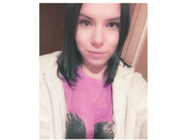 MagdalenaMurchie with black hair, pink shirt, white jacket, and subtle smile, showing her face and upper body in a soft-lit selfie. Her tits are not visible.