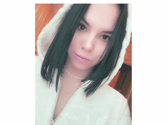MagdalenaMurchie with straight black hair, light skin, and pink lips, wearing a white fluffy robe with a hood, and a delicate silver necklace, looking at the camera.