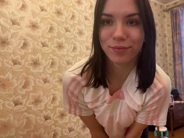 MagdalenaMurchie with dark brown hair, wearing a pink and white striped shirt, leaning forward, smiling slightly, showing her small tits, in a room with floral wallpaper.