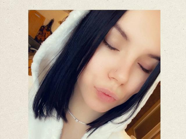 MagdalenaMurchie with closed eyes, black straight hair, white hooded robe, silver necklace, puffy pink lips, pale skin, slight nose, and soft expression. Her ass is not visible.