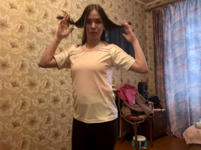 MagdalenaMurchie with brown hair, holding her hair in two hands, wearing a white shirt and dark pants, standing in a room with floral wallpaper, smiling slightly.