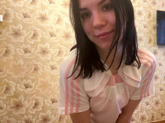 MagdalenaMurchie with shoulder-length black hair, wearing a pink and white striped shirt with a ruffled collar, smiling at the camera, showing her small tits and cute face.