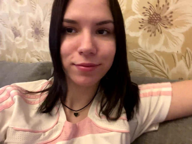 MagdalenaMurchie with medium-length black hair, pink and white shirt, black necklace with heart pendant, slight smile, fair skin, sitting on gray couch, floral-patterned wallpaper in background.