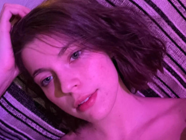 JennellMatacale with short brown hair, blue eyes, light skin, and pinkish lighting, lying down, smiling slightly, one hand in hair, bare shoulders, soft pink and purple hues.