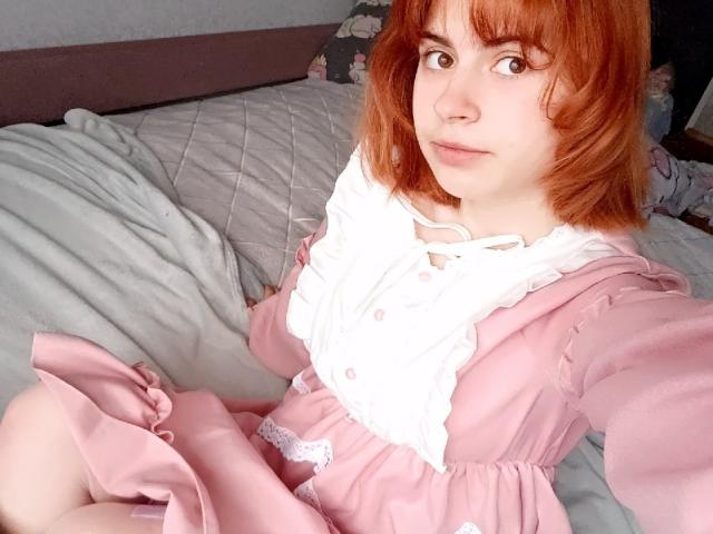 MireilleSpotwood with red hair, wearing a pink dress with white ruffle, sitting on a bed, looking at the camera with a slight smile, showing her pale skin.