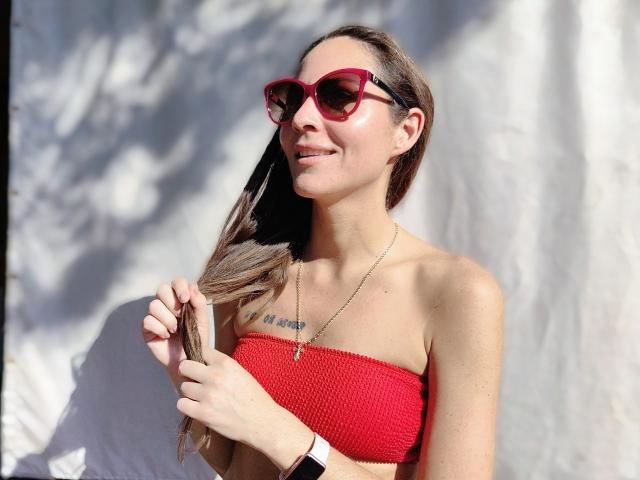 MissKinney with red tube top, holding brown hair, wearing red sunglasses, and a necklace, smiling, showing small tits, wearing a black wristwatch, sunlight on her face.