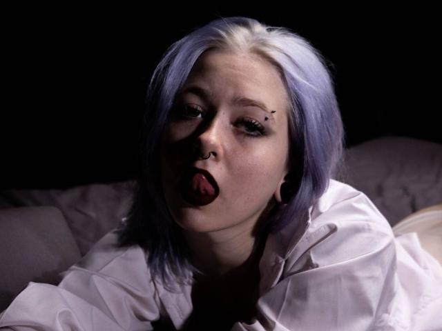 Iris, con capelli viola e bianchi, piercing al naso e eyeliner nero, che mostra la lingua, indossa una camicia bianca, ha degli tappi per le orecchie neri e uno sfondo scuro..