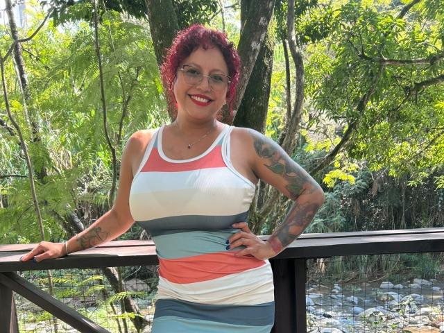 LenaMadisson, con cabello rizado rojo, gafas y tatuajes, vestida con un vestido de tirantes a rayas, de pie en un puente de madera, sonriendo, con las manos en la barandilla, y un fondo de bosque verde..