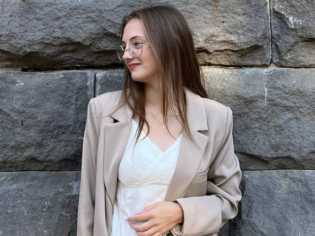 Rosita Appelt mit langen braunen Haaren, Brille, einem beigen Blazer, einem weißen Kleid und den Händen auf dem Bauch, steht vor einer felsigen Wand und zeigt ihre Brüste leicht durch das Kleid hindurch..