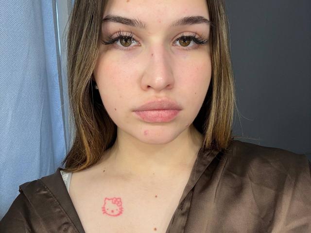 LaylaMantsch with brown hair, fair skin, and a small pink Hello Kitty tattoo on her chest. She's wearing a brown satin shirt, looking straight into the camera with pouty lips.
