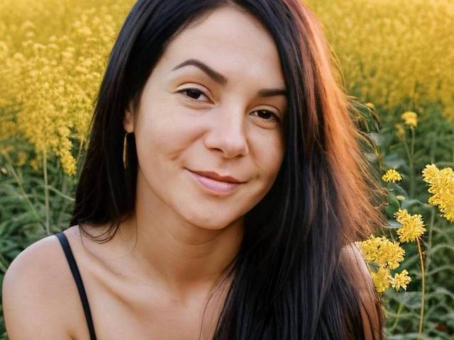 Il viso di SkyNaate, con i suoi lunghi capelli neri, il sorriso appena accennato, gli occhi marroni e il naso piccolo, indossa una canotta nera e si trova in un campo giallo con piante verdi alte..