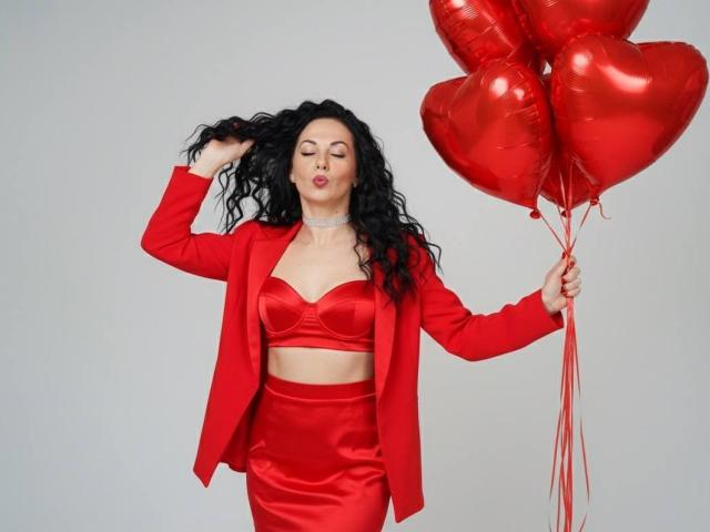 SkyNaate met krullend zwart haar, gekleed in een rode satijnen bh, een bijpassende rok en een blazer, houdt rode hartvormige ballonnen vast, ziet er zelfverzekerd uit, met haar in haar handen, lippen lichtjes opgetrokken..