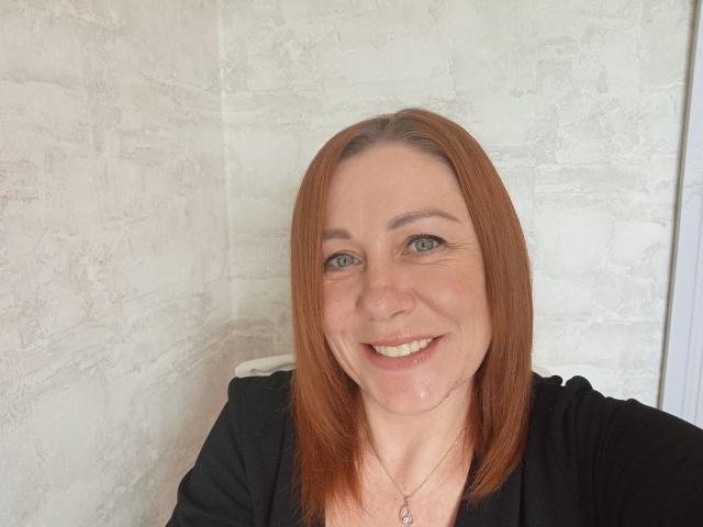 TaylorFoxT has red hair, blue eyes, and a smile showing teeth. She's wearing a black top with a heart necklace, and has a fair skin tone.