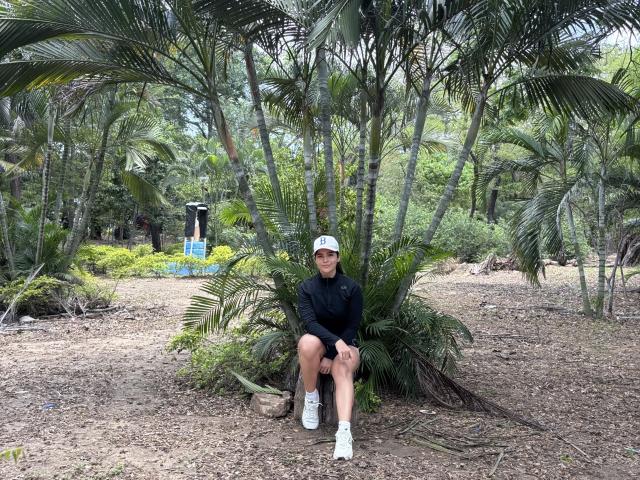 ScarlletZhen is sitting on a tree stump in a forest, wearing a black long-sleeve shirt, white cap, and white sneakers, with a cute, slim, and attractive ass.