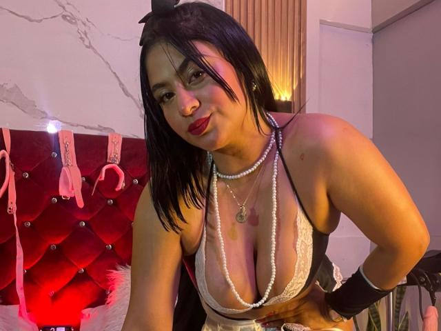 DiannaRosse with black hair, wearing a white lace bra, pearl necklace, black wristband, and red lipstick, leaning forward, showing her large tits, in a dimly lit room.
