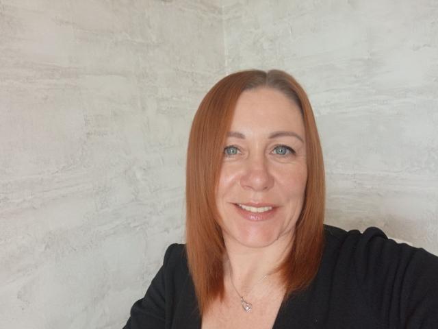 TaylorFoxT has red hair, blue eyes, and a slight smile, wearing a black shirt, with a necklace, looking at the camera, showing her boobs and ass in the photo.