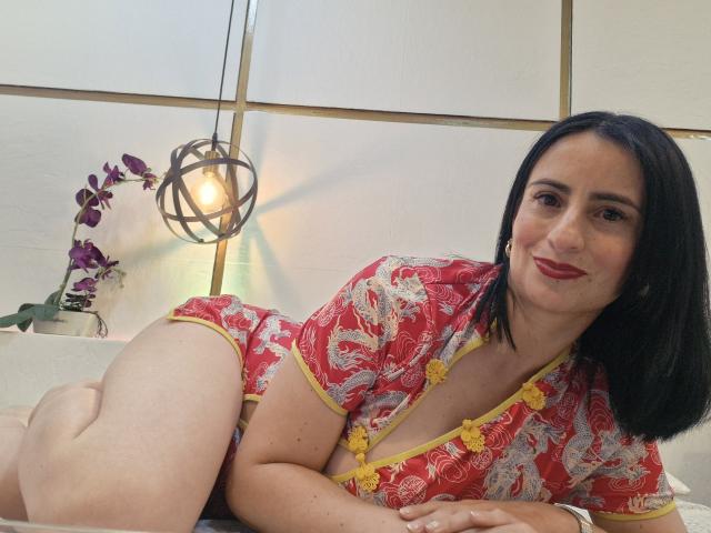 BrendaCantorel, a curvy woman with black hair, red lipstick, and a red floral Chinese dress, lying on a bed, showing her bare ass and cleavage.
