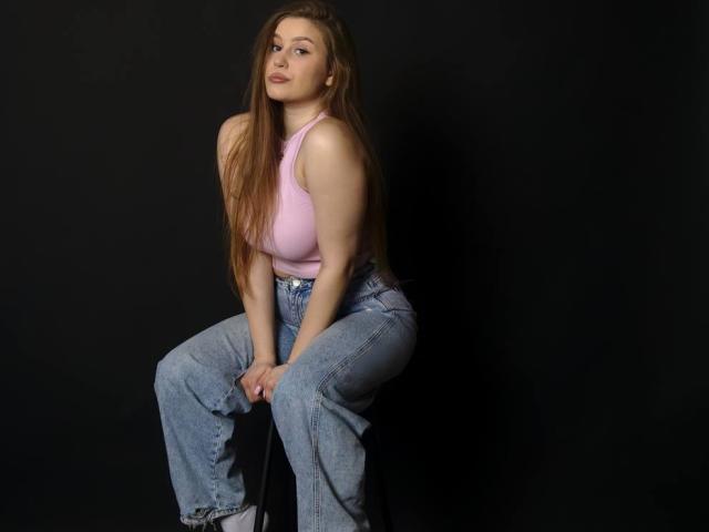Camelia Korman, avec ses longs cheveux bruns, son débardeur rose et son jean bleu taille haute, est assise sur un tabouret devant un fond noir, regardant légèrement sur le côté avec un sourire discret..