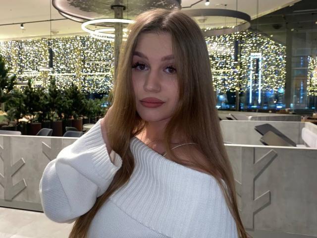 CameliaKorman with long brown hair, wearing a white off-shoulder sweater, has a cute face with pink lipstick and a subtle smile, standing in a well-lit indoor space.
