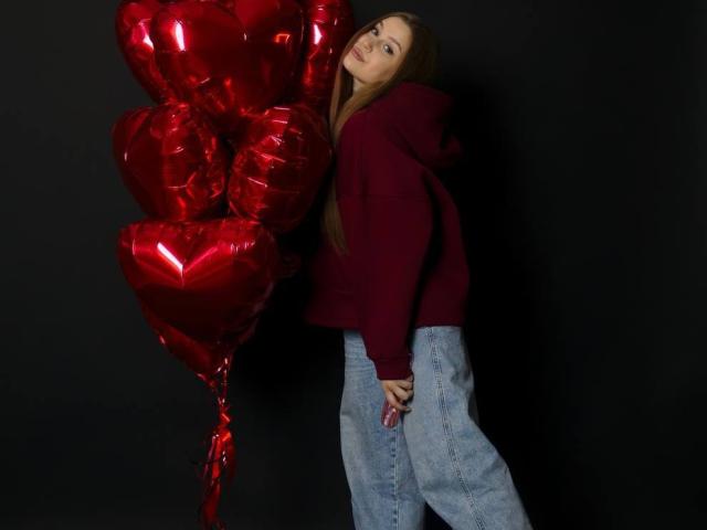 Camelia Korman, com balões vermelhos em formato de coração, vestindo um moletom marrom e jeans azuis, olhando para trás com uma expressão ligeiramente irritada ou entediada..