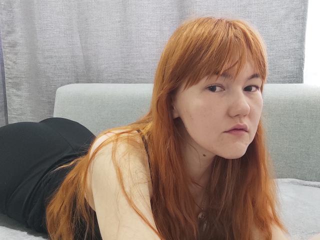 SheritaChieng, with red hair, is lying on a grey couch, wearing a black tank top, showing her pale skin and small, round ass, looking at the camera.