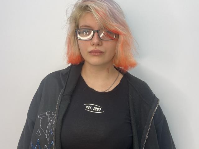 Latisha Rukavina, com cabelo loiro com tons de rosa, óculos pretos, camisa preta, jaqueta preta e colar de prata, olhando diretamente para a câmera com uma expressão neutra..