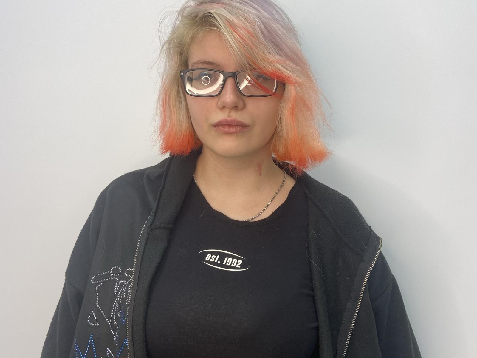 Latisha Rukavina, con i capelli biondi tendenti al rosa, occhiali neri, camicia nera, giacca nera e una collana d'argento, guarda direttamente la fotocamera con un'espressione neutra..