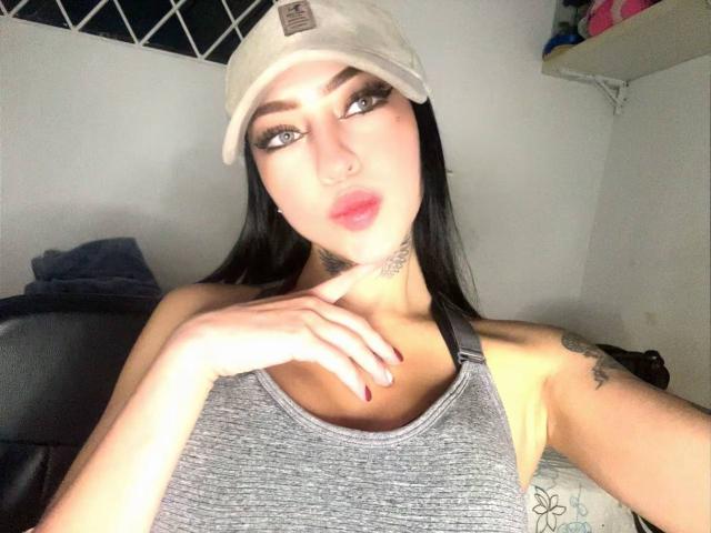 LunaBaster with black hair, wearing a white cap, grey tank top, and red nails, touching her chin, showing tattoos, and looking at the camera. Boobs visible.