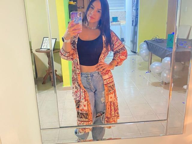 Alina Owens, con lunghi capelli neri, indossa un top corto nero, jeans strappati e un accappatoio colorato con motivi, e sta scattando un selfie allo specchio, mostrando il seno e i glutei..