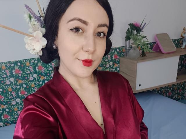 MabyLegrand with black hair, red lipstick, and a shiny red velvet robe, showing her tits, floral headpiece, and a subtle smile. Bedroom with floral wallpaper in the background.