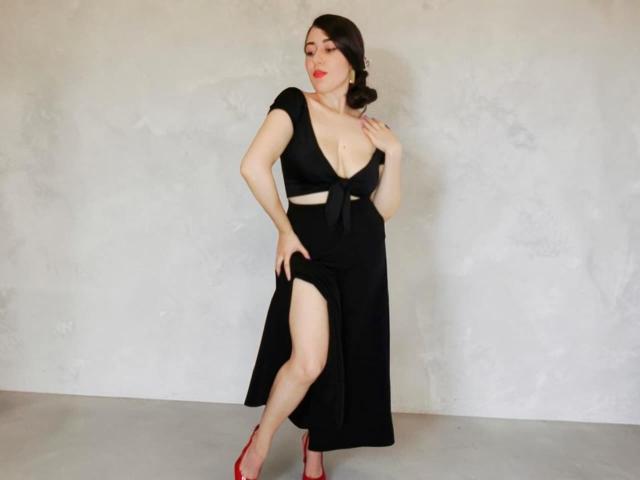 MabyLegrand in a black dress with a deep neckline, showing tits, and a high slit, wearing red heels, standing with one hand on her chest, the other on her thigh.