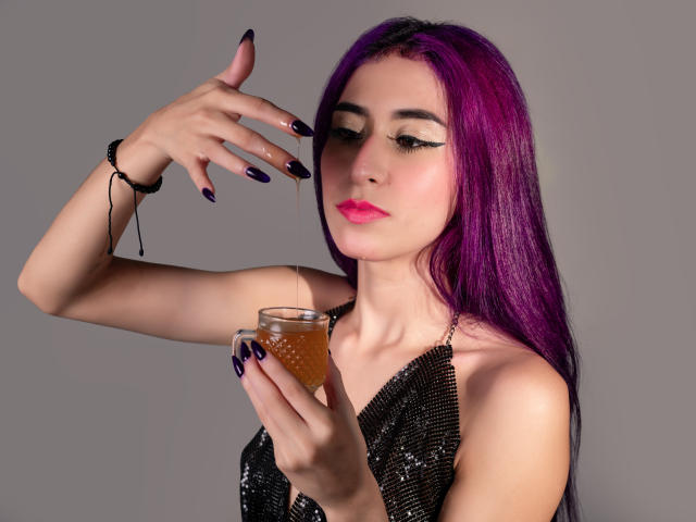 JuliaJobs with purple hair, black eyeliner, and pink lipstick, holding a glass of orange liquid, has dark purple nails, wearing a black sequined dress, and a black bracelet.