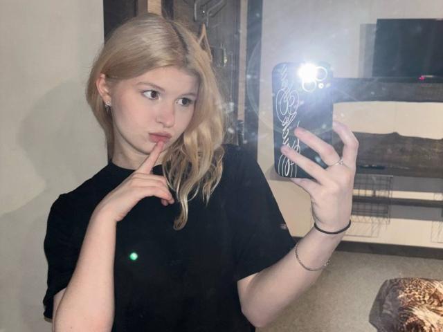 Delphine Hambleton, avec ses cheveux blonds, son t-shirt noir, un doigt sur les lèvres, se prend en selfie devant un miroir avec le flash de l'appareil photo, une bague à la main droite et un bracelet au poignet gauche..