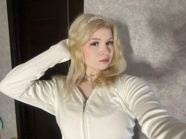 Delphine Hambleton a les cheveux blonds, elle porte un sweat à capuche blanc avec une fermeture éclair, une tiare argentée et un collier ras du cou. Elle a une main dans ses cheveux, mettant ainsi en valeur sa poitrine..