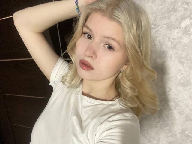 Delphine Hambleton, avec ses cheveux blonds, portant une chemise blanche, avec des lèvres pulpeuses et un bracelet, a son bras levé, mettant en valeur sa petite poitrine et sa peau claire..