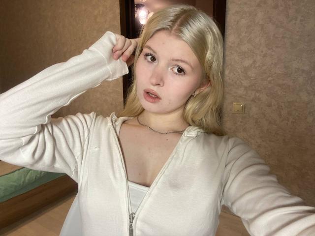 Delphine Hambleton, com cabelo loiro, moletom branco com zíper e maquiagem discreta, segurando o cabelo para cima, mostrando seios pequenos e uma silhueta esguia, em um quarto bem iluminado..