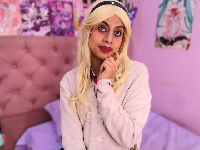 Blond EimyFlorian with pouty red lips, touching chin, wearing white cardigan, purple eyes, pink blush, tufted pink headboard, blonde wig, purple wall, anime posters in background.