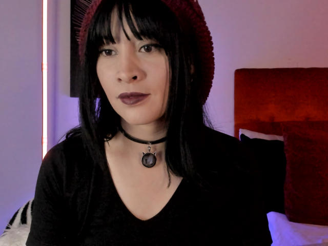 IssisDiosa with black hair, red beanie, black choker with ring, and dark lipstick, wearing a black shirt, in a bedroom with red headboard.