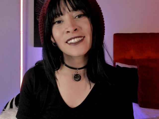 IssisDiosa has black hair, a red beanie, black choker with a metal ring, and a black shirt, smiling with teeth showing. She's wearing small hoop earrings.