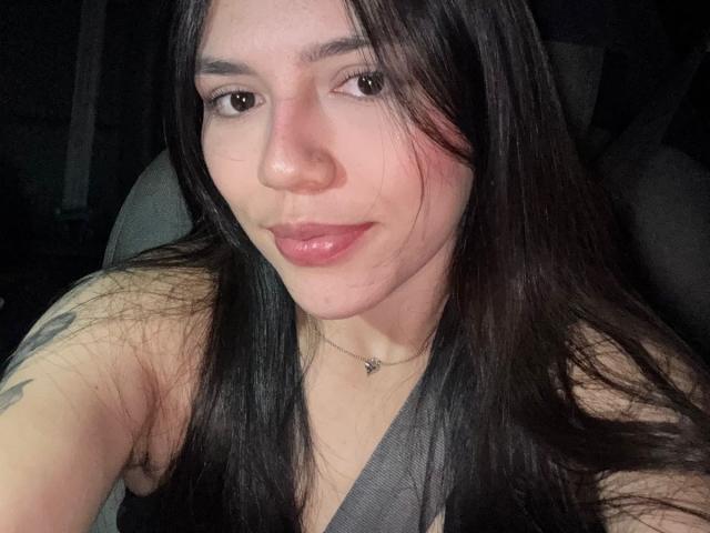 SophiaMarx with long black hair, glossy pink lips, nose ring, and a tattoo on her right arm, wearing a grey seatbelt, smiling softly in a car at night.