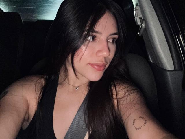 SophiaMarx with dark hair, wearing a black top, seatbelt on, small tattoo on upper arm, sitting in a car at night, looking to the side with a slight smile.