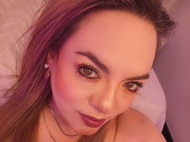 AvaMia's face with pinkish hair, thick eyebrows, brown eyes, dark lipstick, and pink blush, wearing pearl earrings and a necklace, in a soft pink light.