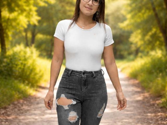 SolaraDubois with small tits, wearing a white t-shirt and ripped, dark grey jeans, standing on a dirt path, has brown hair, glasses, and a subtle smile.