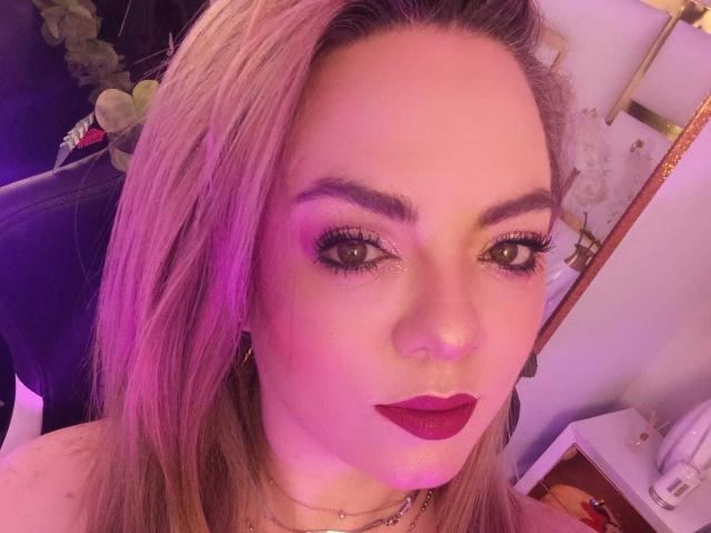 AvaMia with pinkish hair, purple eyelid makeup, dark red lipstick, and a nose ring, looking at the camera with a slight smile. Her tits are partially visible.