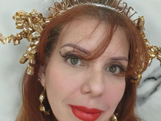 Khharha has red hair, a gold curly headpiece, and is wearing red lipstick with long eyelashes. She has green eyes and is smiling slightly.