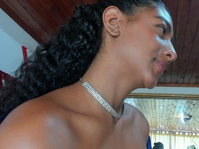 NatalyBlonder, dark-skinned woman with curly black hair in a ponytail, wearing a diamond choker, smiling, showing her nose and ear with a small gold hoop.