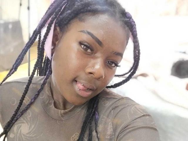 KarinaFlowers with dark skin, purple braided hair, pink ribbon, nose ring, and subtle makeup, wearing a brown patterned top, smiling slightly with a small nose piercing visible.